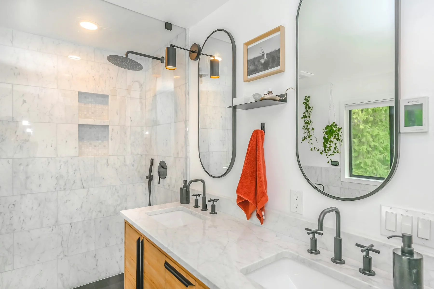 Wide view of marble bathroom showing rain shower head, oval mirrors and oak vanity