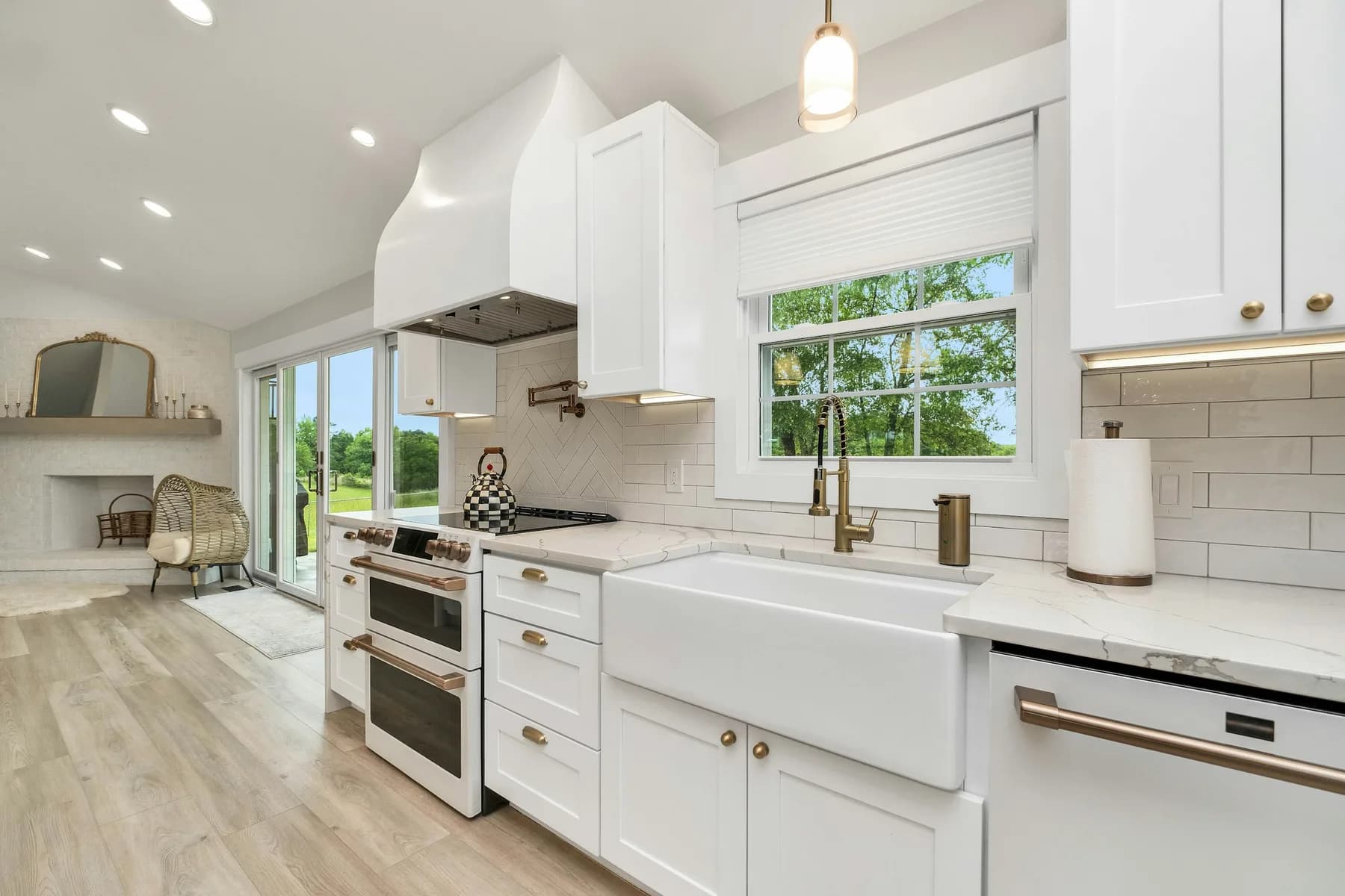 Farmhouse apron sink beneath a window with brass faucet and white shaker cabinets